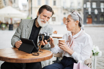 Aged man and woman looking on retro camera while sitting at table with digital tablet. Lovely couple relaxing on cafe terrace and drinking hot coffee.