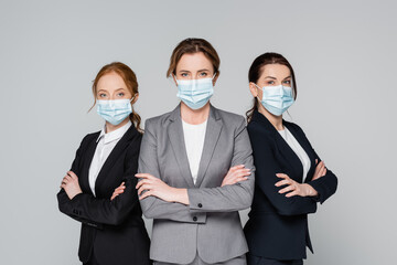 Businesswomen in suits and medical masks looking at camera isolated on grey