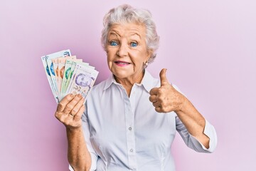 Senior grey-haired woman holding singapore dollars banknotes smiling happy and positive, thumb up doing excellent and approval sign