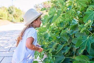Adorable blonde child wearing summer hat smiling happy. Standing with smile on face looking at the leaves around the park
