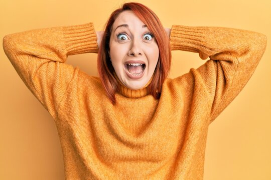 Beautiful redhead woman wearing casual winter sweater over yellow background crazy and scared with hands on head, afraid and surprised of shock with open mouth