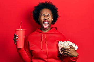 Young african american woman eating popcorn and drinking soda angry and mad screaming frustrated...