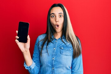 Young hispanic woman holding smartphone showing screen scared and amazed with open mouth for surprise, disbelief face