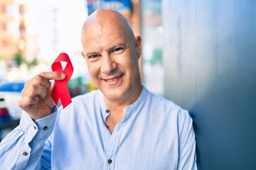 Middle age bald man smiling happy holding red hiv ribbon leaning on the wall at the city