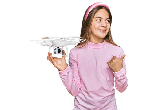 Beautiful brunette little girl using drone pointing thumb up to the side smiling happy with open mouth