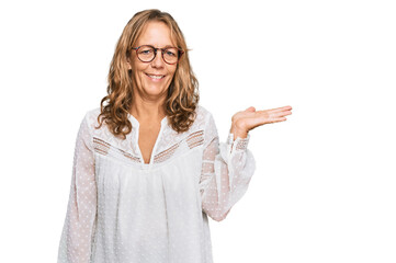 Obraz premium Middle age blonde woman wearing casual white shirt and glasses smiling cheerful presenting and pointing with palm of hand looking at the camera.