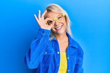 Young blonde girl wearing casual clothes and glasses smiling happy doing ok sign with hand on eye looking through fingers