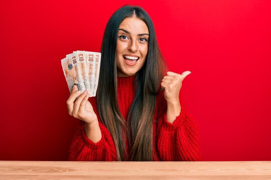 Beautiful hispanic woman holding united kingdom pounds pointing thumb up to the side smiling happy with open mouth