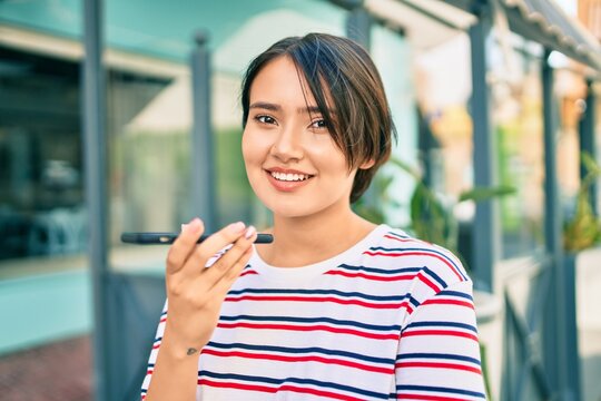 Young latin girl smiling happy sending voice message using smartphone at the city
