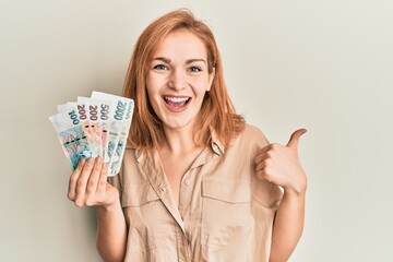 Young caucasian woman holding czech koruna banknotes smiling happy and positive, thumb up doing excellent and approval sign