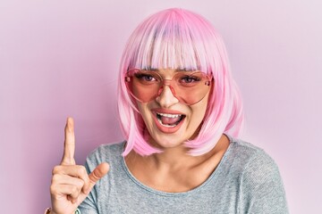 Young caucasian woman wearing pink wig and heart sunglasses smiling with an idea or question pointing finger with happy face, number one