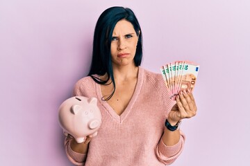 Young caucasian woman holding euro banknotes and piggy bank skeptic and nervous, frowning upset because of problem. negative person.