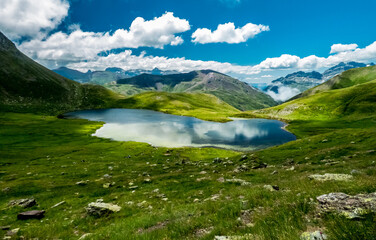 Fototapeta na wymiar Pradera de alta montaña salpicada de rocas que rodean una laguna glaciar, con los altos picos de los Pirineos españoles al fondo