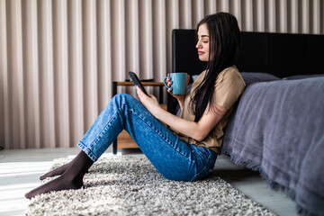Beautiful woman sitting on the floor use phone and drinking coffee. Distance learning online education and remote work.