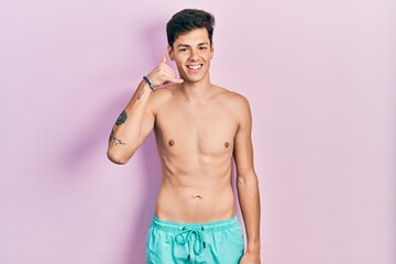 Fototapeta premium Young hispanic man wearing swimwear shirtless smiling doing phone gesture with hand and fingers like talking on the telephone. communicating concepts.
