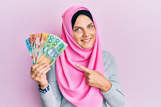Young caucasian woman wearing islamic hijab holding australian dollars banknotes smiling happy pointing with hand and finger