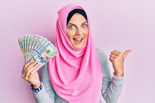 Young caucasian woman wearing islamic hijab holding dollars banknotes pointing thumb up to the side smiling happy with open mouth