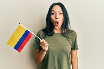 Young hispanic girl holding colombia flag scared and amazed with open mouth for surprise, disbelief face