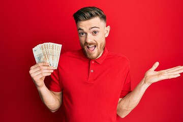 Young redhead man holding 100 danish krone banknotes celebrating achievement with happy smile and winner expression with raised hand