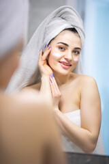 Beauty woman looking in mirror, applying hydrating lotion creme on cheeks, finishing morning domestic skincare routine. Smiling woman grooming herself after showering in bathroom.