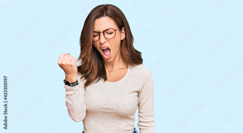 Sticker Young brunette woman wearing casual clothes and glasses angry and mad raising fist frustrated and furious while shouting with anger. rage and aggressive concept.