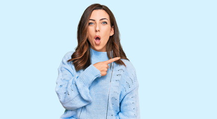 Young brunette woman wearing casual winter sweater surprised pointing with finger to the side, open...