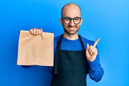 Young hispanic man wearing waiter uniform holding take away paper bag smiling happy pointing with hand and finger to the side