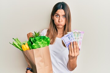 Brunette young woman holding groceries and indian rupee banknotes puffing cheeks with funny face....
