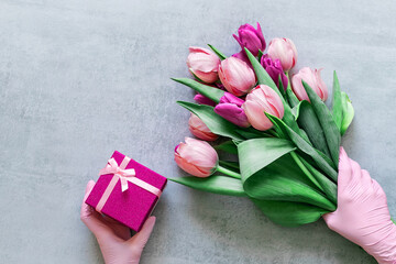 Women's hands in pink medical gloves hold a bouquet of tulips and a gift box on a gray background. Gift