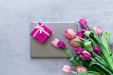 A gray laptop, a pink gift box, and a bouquet of tulips on a gray concrete background. Gift