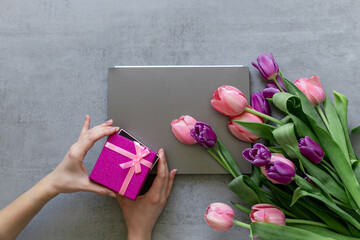 A woman's hands open a pink gift box. A gray laptop and pink tulips on a gray concrete background.