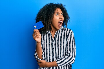 Beautiful african american woman with afro hair holding credit card angry and mad screaming frustrated and furious, shouting with anger. rage and aggressive concept.