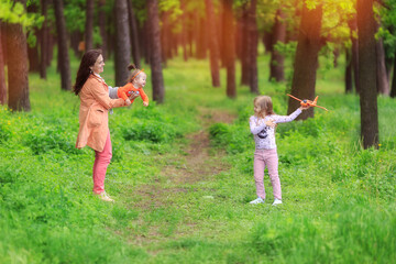 Naklejka na ściany i meble Happy mother with two dauthers playing in park. summer day. Woman throwing baby up in air. Girl holds in hands model of aircraft. Family have fun with toy airplanes outdoors. Happy childhood vacation