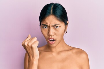 Young chinese woman standing topless showing skin angry and mad raising fist frustrated and furious...