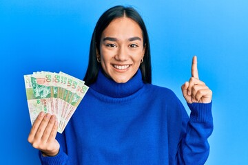 Young asian woman holding 50 hong kong dollars banknotes smiling with an idea or question pointing finger with happy face, number one