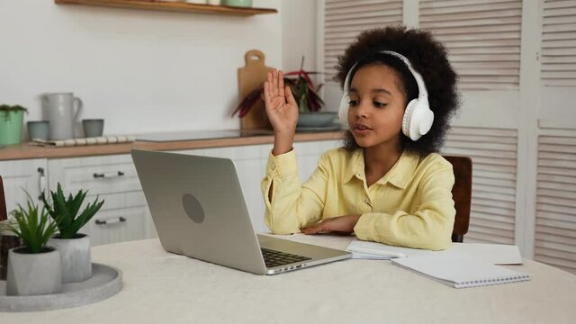Little African American girl in headphones uses laptop to video call with teacher. Online lecture, girl raises hand and answers questions. Teenage is sitting at table in kitchen. Slow motion.