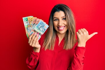 Beautiful brunette woman holding australian dollars pointing to the back behind with hand and thumbs up, smiling confident