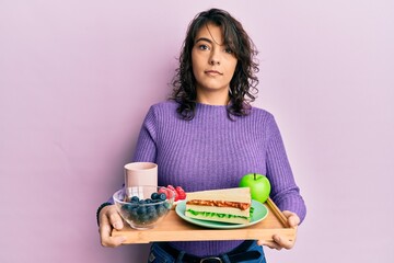 Young hispanic woman holding tray with breakfast food relaxed with serious expression on face. simple and natural looking at the camera.