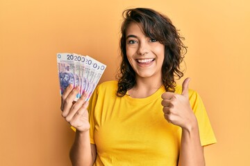 Young hispanic woman holding 20 polish zloty banknotes smiling happy and positive, thumb up doing...