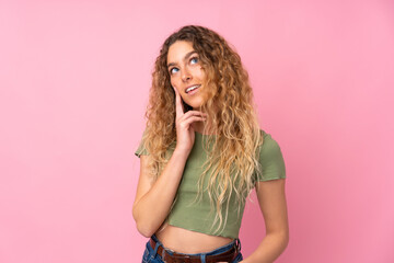 Young blonde woman with curly hair isolated on pink background thinking an idea while looking up
