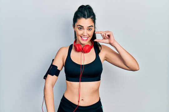 Young hispanic girl wearing gym clothes and using headphones smiling and confident gesturing with hand doing small size sign with fingers looking and the camera. measure concept.