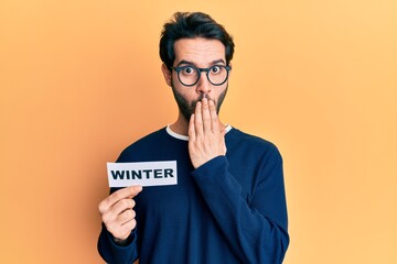 Young hispanic man wearing glasses holding winter word on paper covering mouth with hand, shocked...