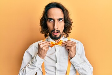 Young hispanic man wearing hipster elegant look holding bowtie in shock face, looking skeptical and...