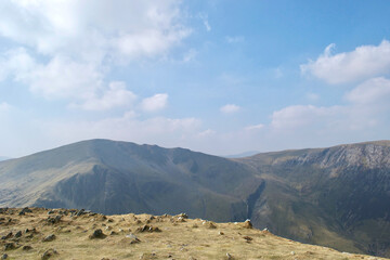 Mountain tops; looking over a rock strewn grassy summit to further peaks in the distance beneath a cloudy blue sky