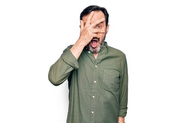 Middle age handsome man wearing casual green shirt over isolated white background peeking in shock covering face and eyes with hand, looking through fingers afraid