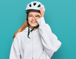 Beautiful young caucasian girl wearing bike helmet smiling happy doing ok sign with hand on eye looking through fingers