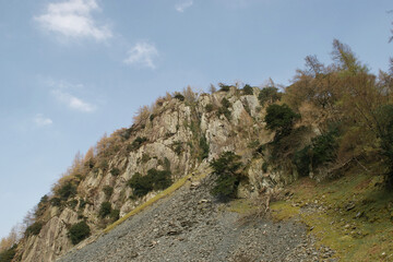 Crags at the rocky peak of a mountain; slag heaps from slate mining, green pines and bare winter trees. Blue sky above the wooded summit. English Lake District, Cumbria, UK