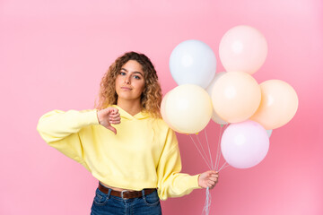 Young blonde woman with curly hair catching many balloons isolated on pink background showing thumb...