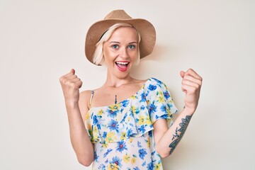 Young blonde woman with tattoo wearing summer hat screaming proud, celebrating victory and success very excited with raised arms