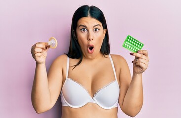 Beautiful brunette woman holding condom and birth control pills in shock face, looking skeptical...